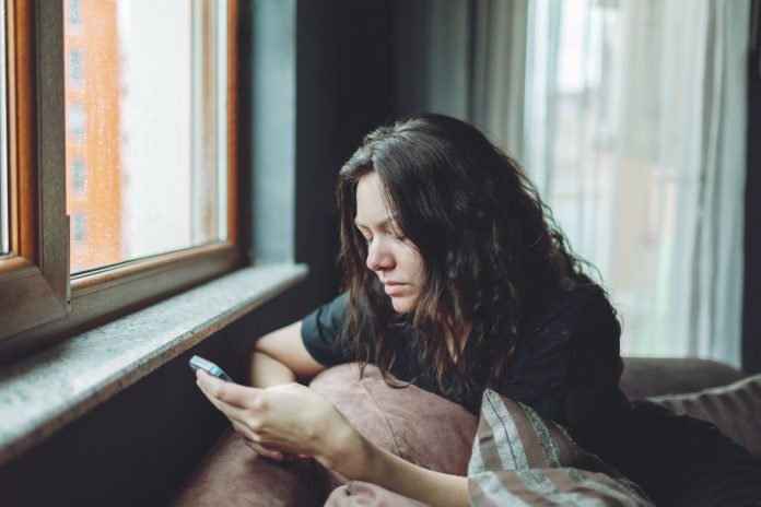 Depressed woman sitting on sofa at home with mobile