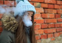 Vape teenager. Young pretty white girl in blue cap and green jacket smoking an electronic cigarette opposite brick wall on the street in the spring.