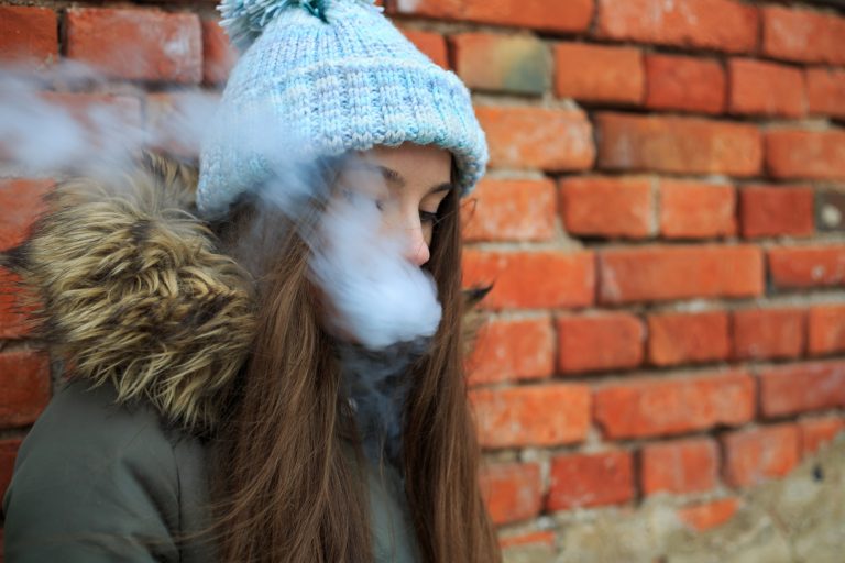 Vape teenager. Young pretty white girl in blue cap and green jacket smoking an electronic cigarette opposite brick wall on the street in the spring.