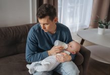 Unhappy young father feeding newborn baby with milk bottle on couch at home. Depressed single dad tired from sleepless night, fatherhood, take care of infant sun. Paternity and paternity leave concept