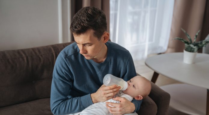 Unhappy young father feeding newborn baby with milk bottle on couch at home. Depressed single dad tired from sleepless night, fatherhood, take care of infant sun. Paternity and paternity leave concept