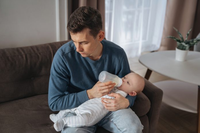 Unhappy young father feeding newborn baby with milk bottle on couch at home. Depressed single dad tired from sleepless night, fatherhood, take care of infant sun. Paternity and paternity leave concept