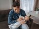 Unhappy young father feeding newborn baby with milk bottle on couch at home. Depressed single dad tired from sleepless night, fatherhood, take care of infant sun. Paternity and paternity leave concept