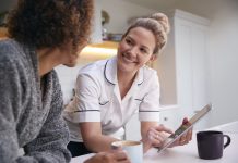 Mature Woman In Dressing Gown At Home Talking With Female Nurse With Digital Tablet