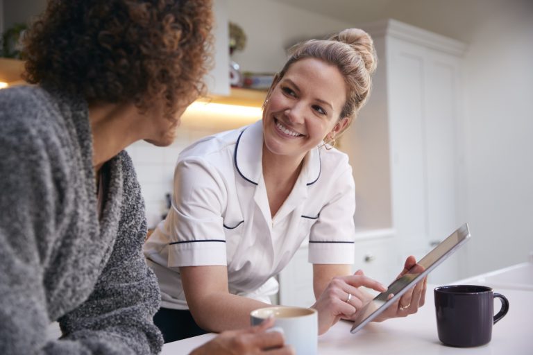 Mature Woman In Dressing Gown At Home Talking With Female Nurse With Digital Tablet