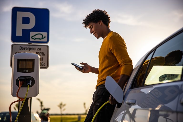 African american man using mobile phone while he is waiting for his electric car to be charged at charging station