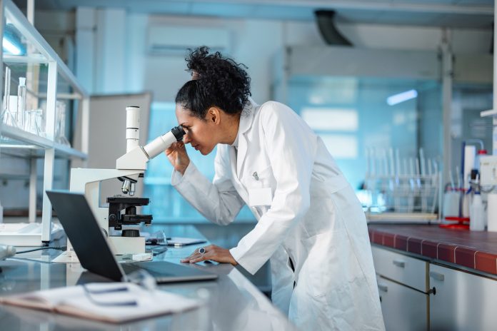 Young Hispanic scientist wearing a lab coat, looking under microscope while using laptop in a laboratory.