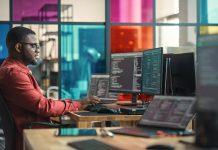 African American Man Writing Lines Of Code On Desktop Computer With Multiple Monitors and Laptop in Creative Office. Male Data Scientist Working on Innovative Online Service For Start-up Company.