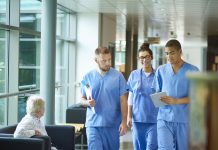 three junior doctors walking along a hospital corridor discussing case and wearing scrubs. A patient or visitor is sitting in the corridor as they walk past .