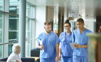 three junior doctors walking along a hospital corridor discussing case and wearing scrubs. A patient or visitor is sitting in the corridor as they walk past .