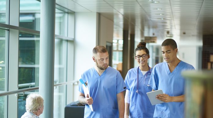 three junior doctors walking along a hospital corridor discussing case and wearing scrubs. A patient or visitor is sitting in the corridor as they walk past .