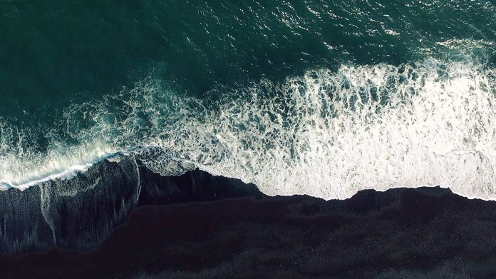 Cold ocean waves washing down the black sand beach making a lot of ocean foam. The video is taken by drone which goes down to 350 meters and rotates.