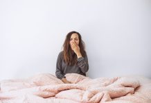 The girl is in bed and yawns. She covers her mouth with her hand. Early morning and she is sleepy.