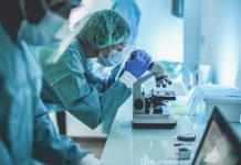 Scentist people working inside laboratory with microscope and laptop while wearing hazmat suit and face masks - Focus on the young woman with microscope