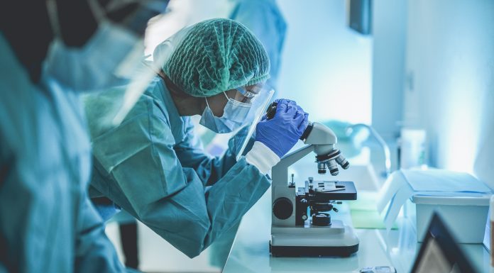 Scentist people working inside laboratory with microscope and laptop while wearing hazmat suit and face masks - Focus on the young woman with microscope