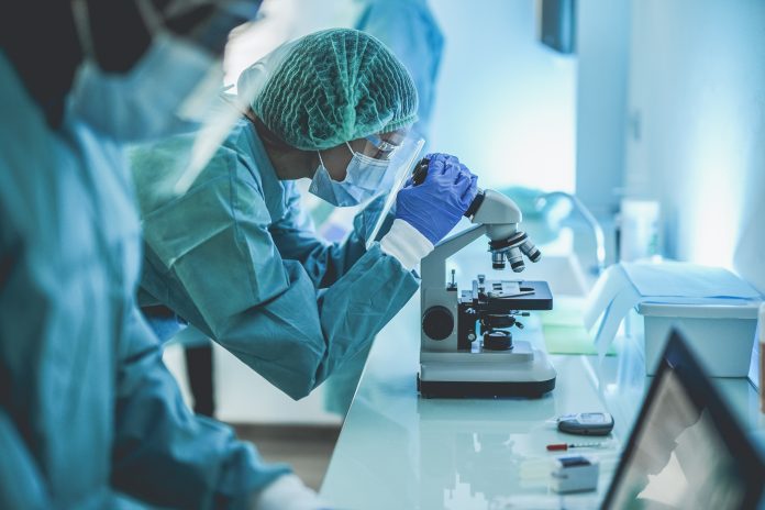 Scentist people working inside laboratory with microscope and laptop while wearing hazmat suit and face masks - Focus on the young woman with microscope