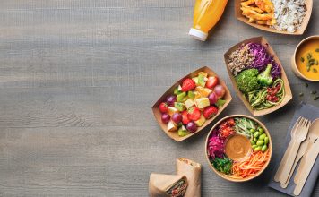 Healthy take away food and drink in disposable eco friendly paper containers on gray wooden background, top view. Fresh salad, soup, poke bowl, vegetable, fruits, chicken wrap and juice.