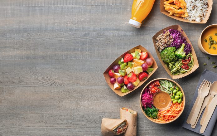 Healthy take away food and drink in disposable eco friendly paper containers on gray wooden background, top view. Fresh salad, soup, poke bowl, vegetable, fruits, chicken wrap and juice.