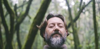 Front portrait of mature man with beard and closed eyes in outdoor leisure activity alone. Male people in meditation with green nature background. Environment and alternative adventure journey