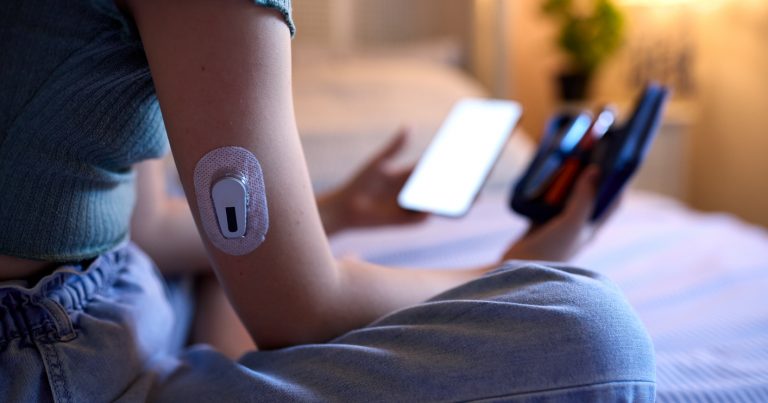 Close Up Of Diabetic Girl Sitting On Bed At Home Checking Insulin Levels On Mobile Phone App