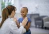 A female Caucasian doctor and a young girl of African descent are indoors in a hospital room. The girl has cancer. She is smiling and giving a high-five to her doctor.