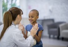 A female Caucasian doctor and a young girl of African descent are indoors in a hospital room. The girl has cancer. She is smiling and giving a high-five to her doctor.