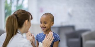 A female Caucasian doctor and a young girl of African descent are indoors in a hospital room. The girl has cancer. She is smiling and giving a high-five to her doctor.