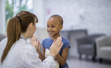 A female Caucasian doctor and a young girl of African descent are indoors in a hospital room. The girl has cancer. She is smiling and giving a high-five to her doctor.