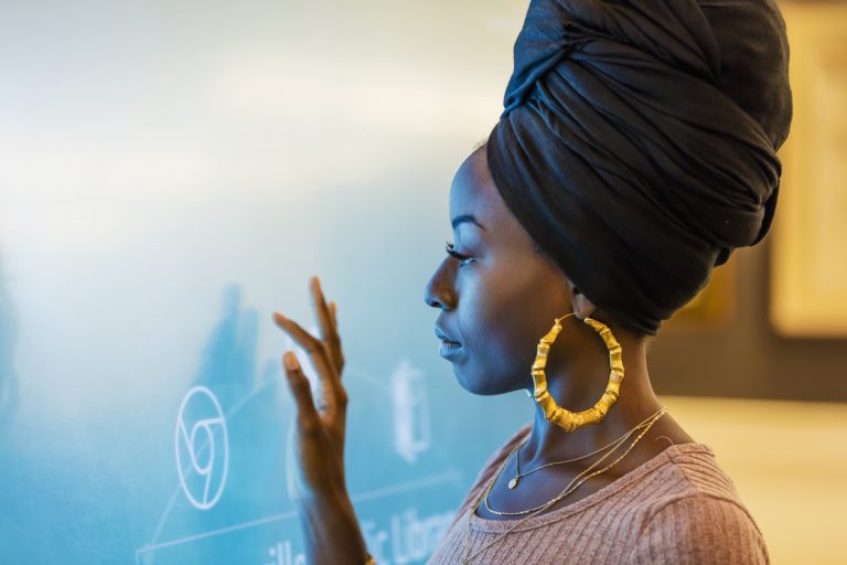 A young African-American woman in her 20s wearing a turban and large earrings, touching a giant interactive display screen.