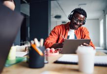 Happy African-American student sitting at desk with his unrecognizable friend while listening online lesson on his digital tablet and writing homework in a notebook.