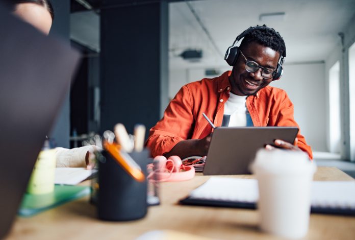 Happy African-American student sitting at desk with his unrecognizable friend while listening online lesson on his digital tablet and writing homework in a notebook.