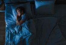 Top View of Handsome Young Man Sleeping Cozily on a Bed in His Bedroom at Night. Blue Nightly Colors with Cold Weak Lamppost Light Shining Through the Window.