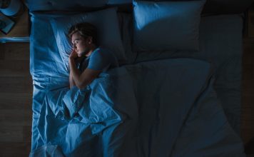 Top View of Handsome Young Man Sleeping Cozily on a Bed in His Bedroom at Night. Blue Nightly Colors with Cold Weak Lamppost Light Shining Through the Window.