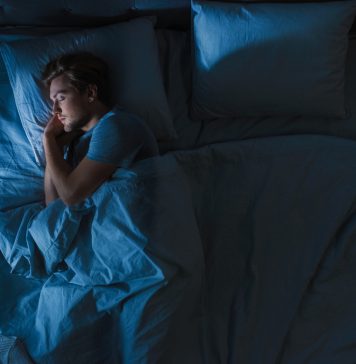 Top View of Handsome Young Man Sleeping Cozily on a Bed in His Bedroom at Night. Blue Nightly Colors with Cold Weak Lamppost Light Shining Through the Window.