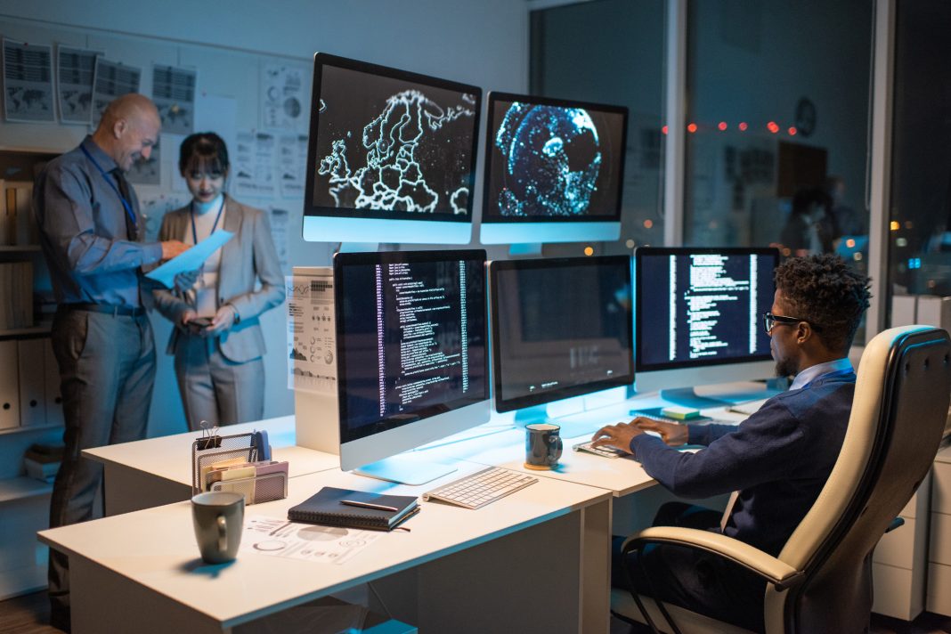 Young African businessman looking at data on computer screen while sitting in armchair in front of several monitors and typing
