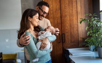 Portrait of young happy man and woman holding newborn cute babe dressed in white unisex clothing.