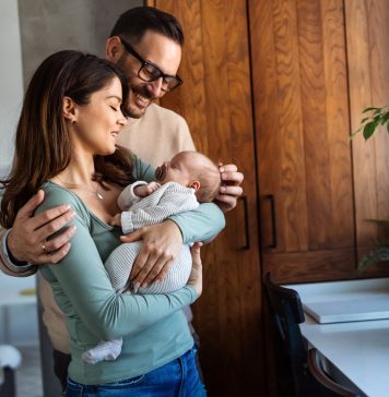 Portrait of young happy man and woman holding newborn cute babe dressed in white unisex clothing.