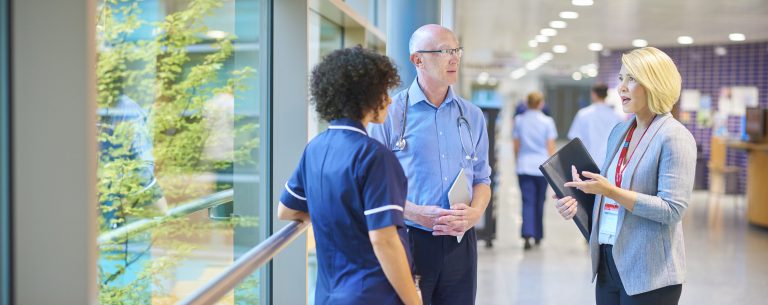 a business woman chats to a doctor and senior staff nurse in a busy hospital corridor