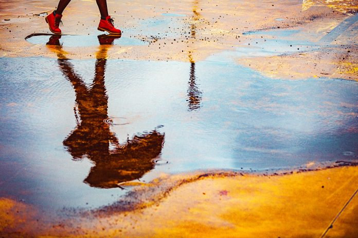 Reflection in a puddle of a young woman holding umbrella, walking outdoors over an empty yellow parking lot on a cold, windy, rainy autumn day. Walking in the rain