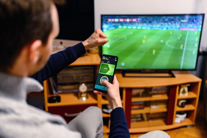 Young men sitting on the sofa at home and watching soccer game. He is using smart phone for sports betting.