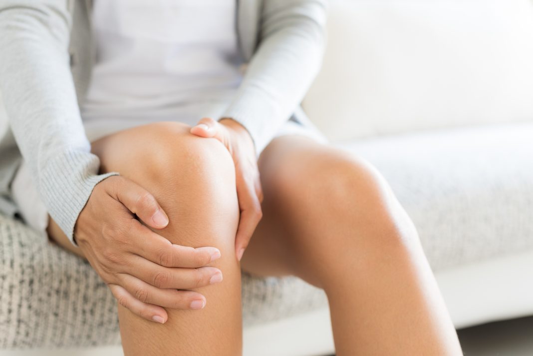 Closeup young woman sitting on sofa and feeling knee pain and she massage her knee at home. Healthcare and medical concept.