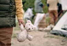 Hand of homeless child holding white teddybear while standing against refugee camp