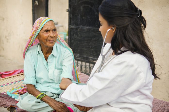 Female doctor examining indian senior adult woman with stethoscope during house visit in.
