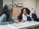 Cheerful instructor in early 50s sitting at desk next to multiracial student, talking and smiling as they discuss her writing assignment in secondary classroom.