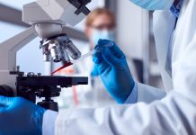 Two Female Lab Workers Wearing PPE Testing Blood Samples In Laboratory With Microscope