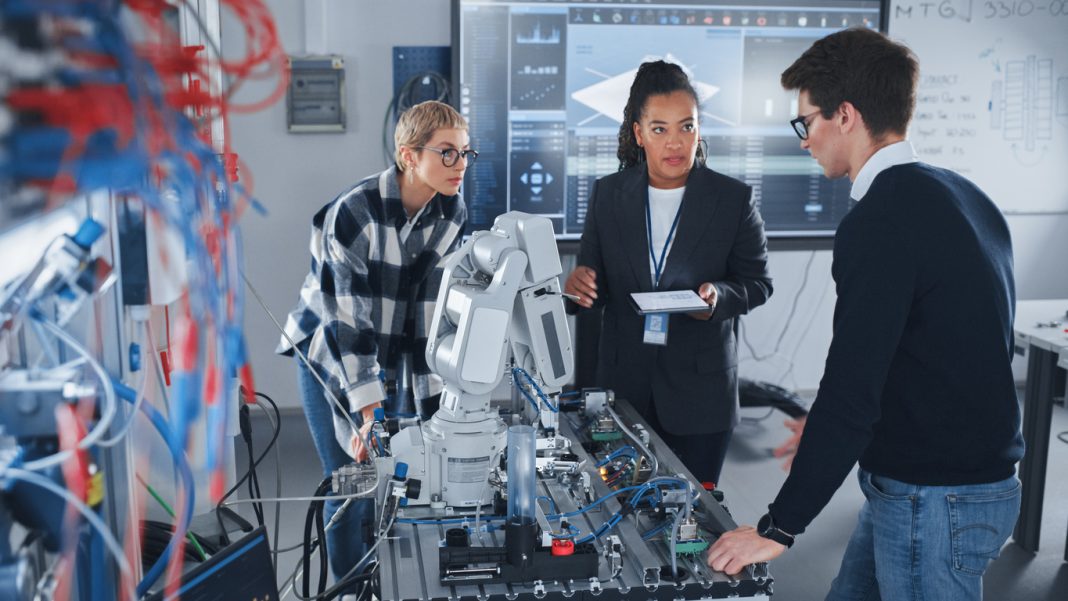 In Robotics Development Laboratory: Black Female Teacher and Two Students Work With Prototype of Robotic Hand. Young Student Telling Something with Smile and Brainstorming with Her Team.