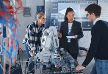 In Robotics Development Laboratory: Black Female Teacher and Two Students Work With Prototype of Robotic Hand. Young Student Telling Something with Smile and Brainstorming with Her Team.