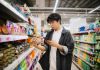 Asian Chinese Student Looking At Smartphone While Choosing Items In Supermarket.