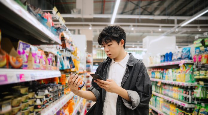 Asian Chinese Student Looking At Smartphone While Choosing Items In Supermarket.