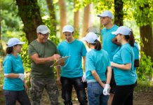 Group of people wearing matching hats and t-shirts, cleaning up the park gathered around the senior man organizing them, holding the notepad